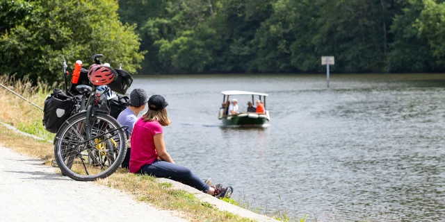 Un couple de cycliste est assis le long de la rivière. Ils observent un petit bateau au deuxième plan.