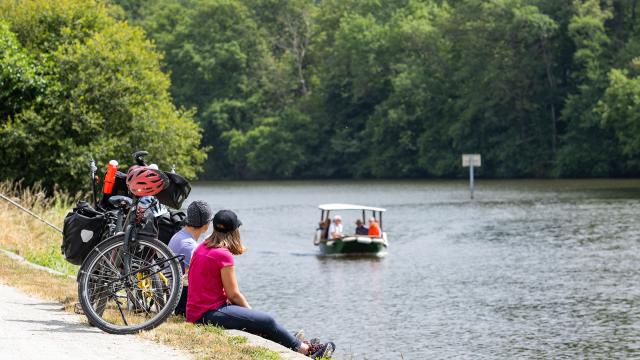 Un couple de cycliste est assis le long de la rivière. Ils observent un petit bateau au deuxième plan.