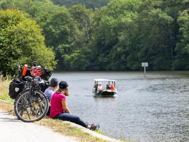 Un couple de cycliste est assis le long de la rivière. Ils observent un petit bateau au deuxième plan.