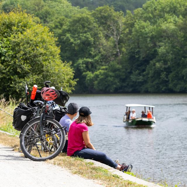 Un couple de cycliste est assis le long de la rivière. Ils observent un petit bateau au deuxième plan.