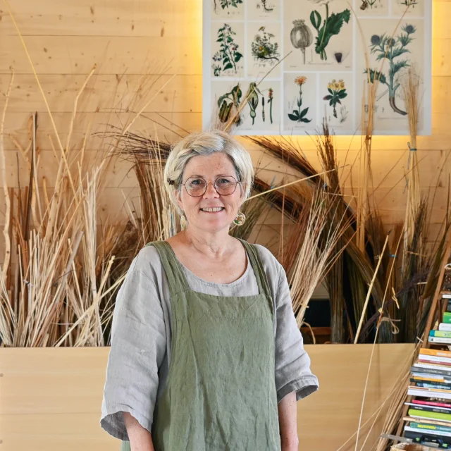 Judith Charpentier de face dans son atelier. Derrierre elle, au mur on voit une affiche qui présente différents végétaux un peu comme les vieilles affiches des écoles d'antan. On voit également différentes tiges d'osier.