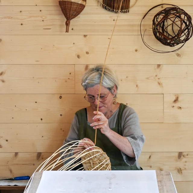 Judith Charpentier est assise à une table. Elle est en plein travail de tressage de l'osier