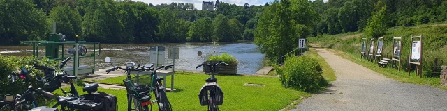 Des vélos sont garés à l'écluse de la Benâtre. En arrière plan on voit la rivière la Mayenne et au fond le château de la Roche domine la vallée verdoyante