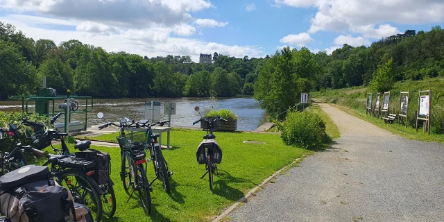 Des vélos sont garés à l'écluse de la Benâtre. En arrière plan on voit la rivière la Mayenne et au fond le château de la Roche domine la vallée verdoyante