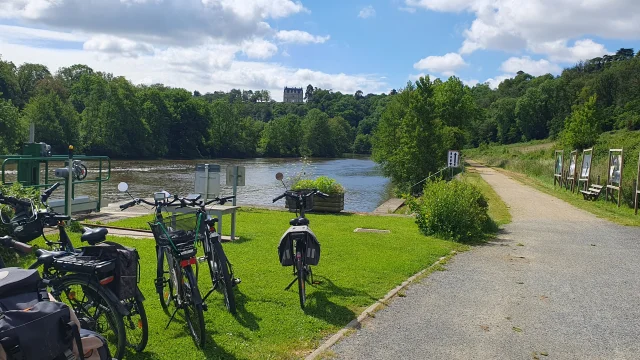 Des vélos sont garés à l'écluse de la Benâtre. En arrière plan on voit la rivière la Mayenne et au fond le château de la Roche domine la vallée verdoyante