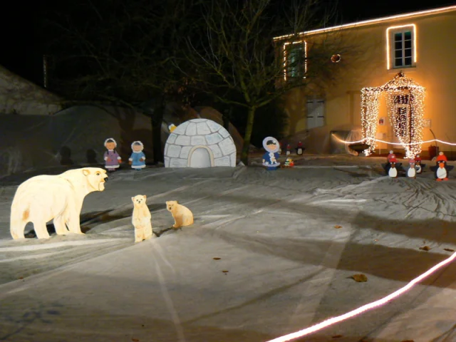 Illuminations de Noël à Athée. On y voit dans le fond une maison avec des guirlande lumineuse. La pelouse a été recouverte d'un tissu blanc pour donner l'illusion de la neige. Un décor présente un igloo, une ourse polaire et deux oursons