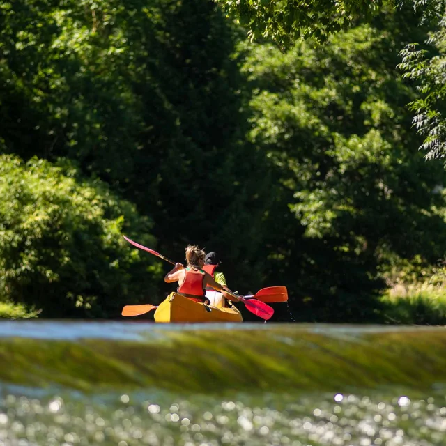 On voit deux personnes de dos naviguant sur un canoë. Les berges de la rivière sont arborées