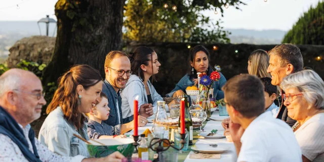 Une grande famille est attablée en extérieur. La lumière douce évoque un début de soirée en été. La table est joliment dressée avec de grandes bougies. Les personnages sont souriant et semblent passer un bon moment. A l'arrière plan on distingue la campagne mayennaise.