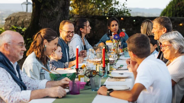 Une grande famille est attablée en extérieur. La lumière douce évoque un début de soirée en été. La table est joliment dressée avec de grandes bougies. Les personnages sont souriant et semblent passer un bon moment. A l'arrière plan on distingue la campagne mayennaise.