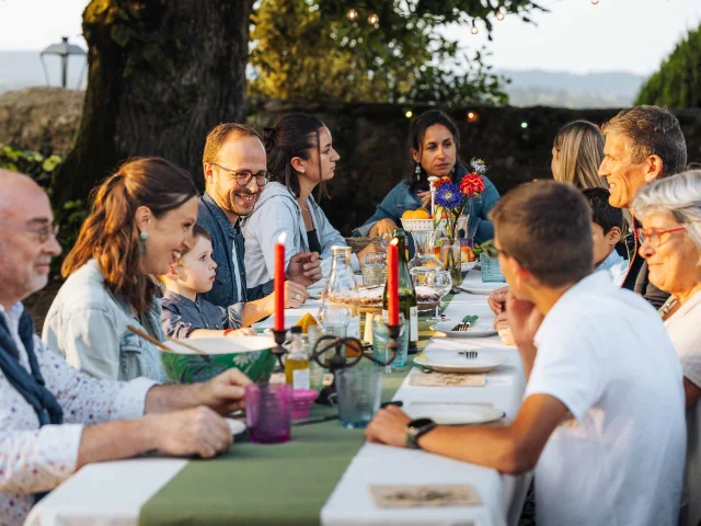 Une grande famille est attablée en extérieur. La lumière douce évoque un début de soirée en été. La table est joliment dressée avec de grandes bougies. Les personnages sont souriant et semblent passer un bon moment. A l'arrière plan on distingue la campagne mayennaise.