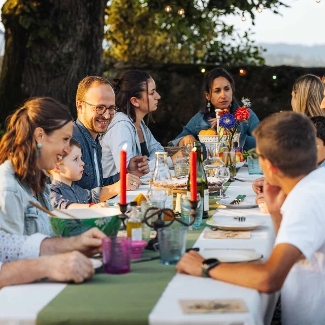 Une grande famille est attablée en extérieur. La lumière douce évoque un début de soirée en été. La table est joliment dressée avec de grandes bougies. Les personnages sont souriant et semblent passer un bon moment. A l'arrière plan on distingue la campagne mayennaise.